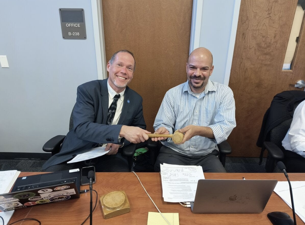 a white man on the left holds a gavel at a desk while a Black man sitting on his right holds the other side of the gavel in a handoff and they are both smiling 